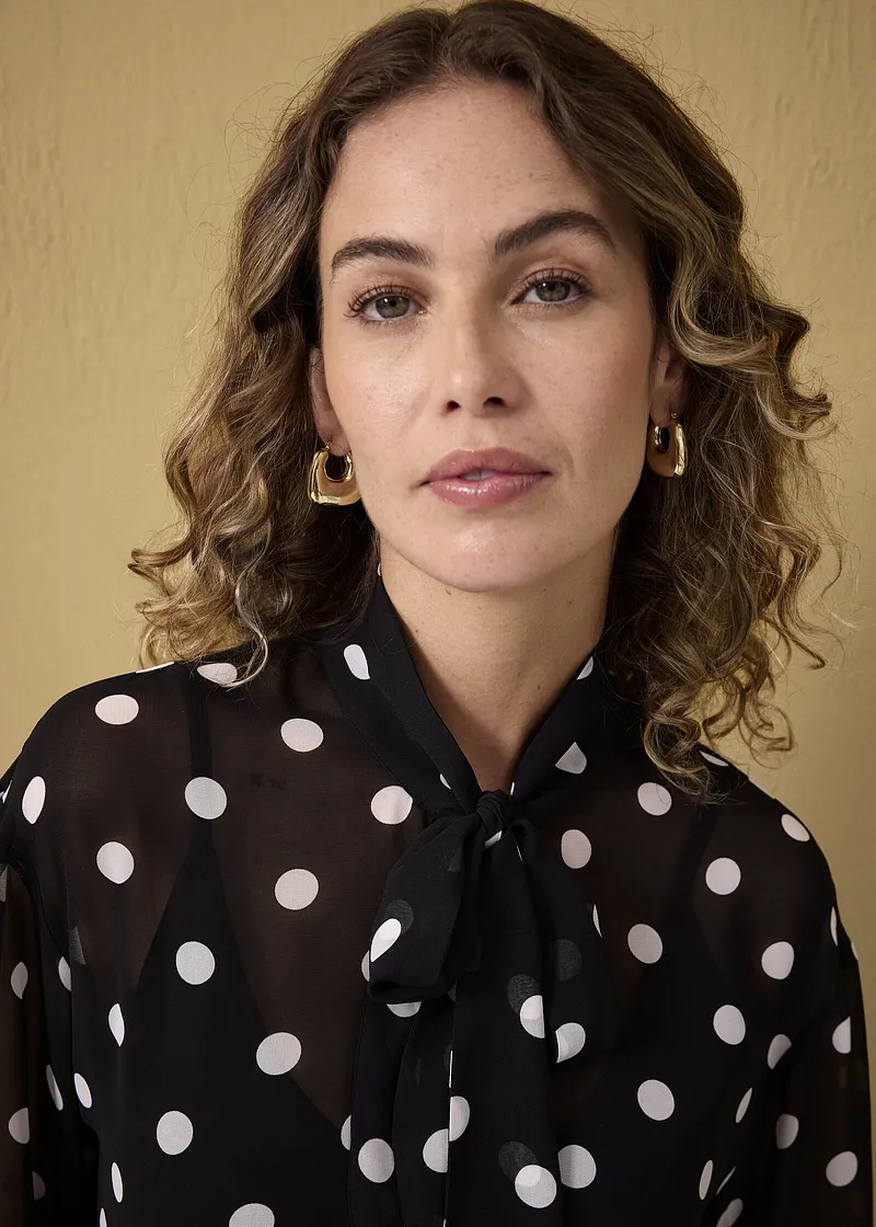 Chemisier élégant noir à pois blancs avec nœud au col. Des boucles d'oreilles de couleur dorée finalisent le look.
