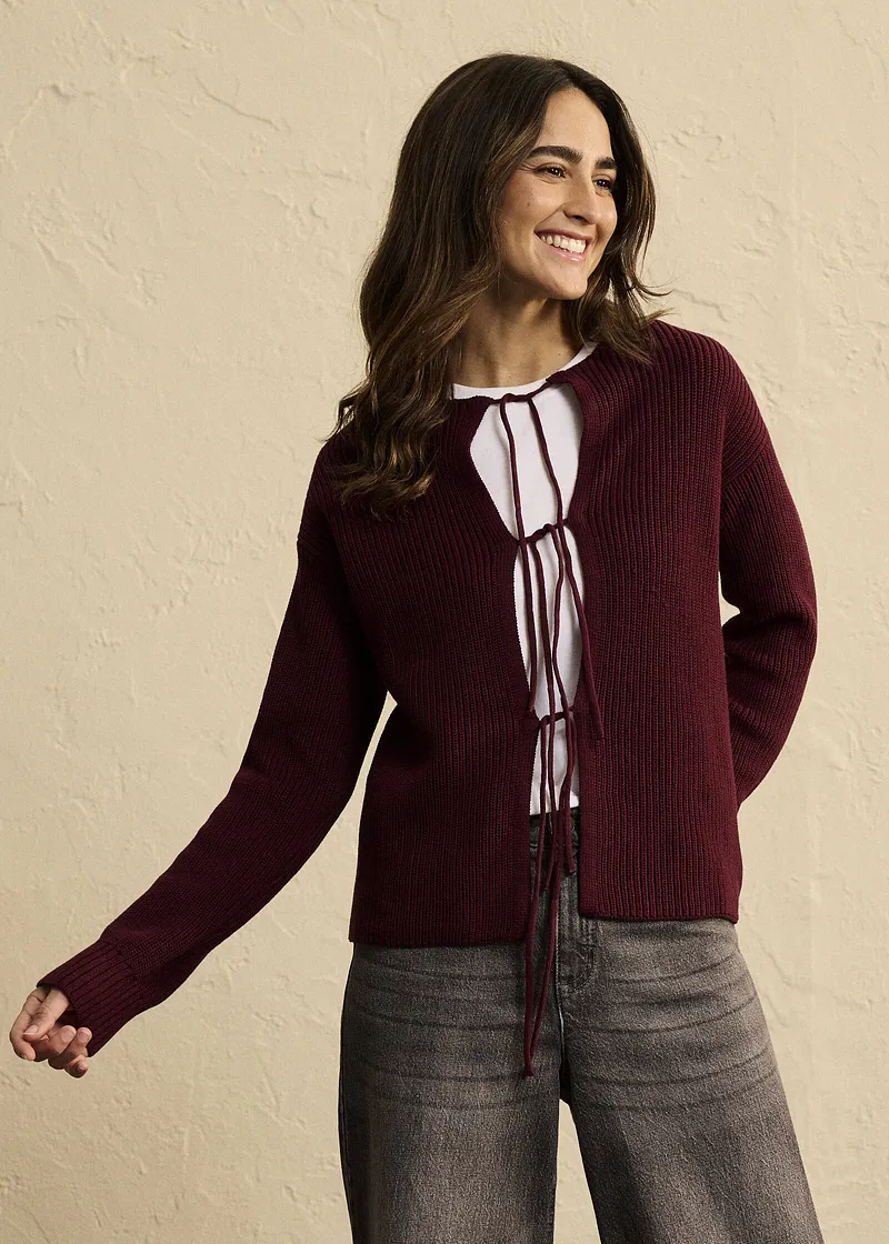 Association d'un gilet en maille rouge bordeaux à laçages, un haut blanc et un pantalon gris.