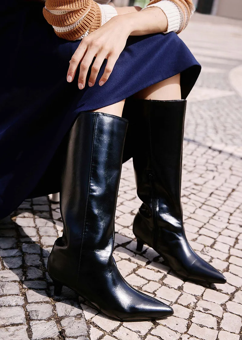 Contraste chic : pull beige, jupe bleu foncé et bottes noires vernies sur une terrasse pavée.