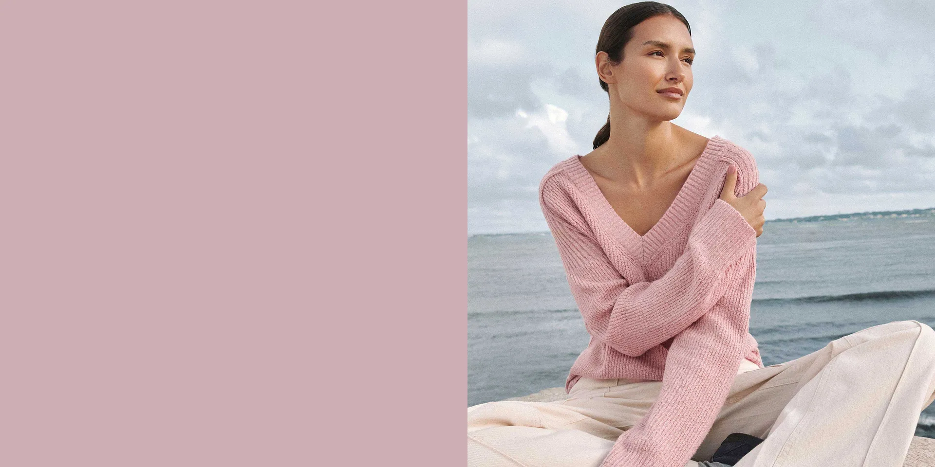Un pull en maille rose tendre avec encolure en V et la mer pour décor de fond.