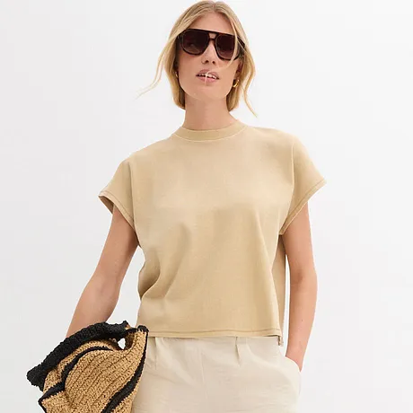 Tenue estivale avec haut beige à manches courtes, pantalon blanc, lunettes de soleil et sac en paille.