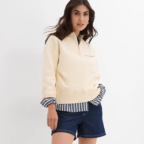 Pull couleur crème par dessus un chemisier rayé, associé à un short en jean et des sneakers blanches.