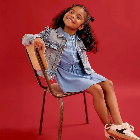 Enfant joyeuse en robe en jean et veste en jean, assise sur une chaise sur fond rouge.