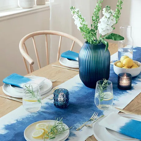 Un chemin de table bleu décore la table de salle à manger, en contraste avec des assiettes blanches et des accessoires colorés.