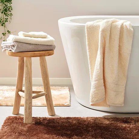 Des serviettes de toilette douces en couleur crème et tons de beige sont posées sur la baignoire et sur un tabouret dans une salle de bain claire.