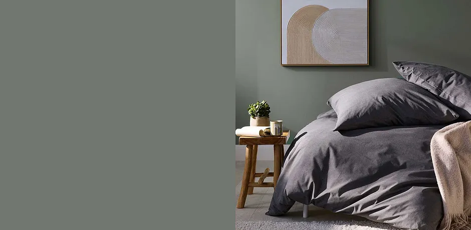 Une élégante parure de lit grise s’accorde avec un mur vert. Une plante complète l’ambiance sobre de cette chambre.